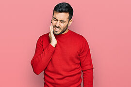 jeune homme en pull rouge sur un fond rouge clair se tenant la joue avec une expression de douleur, les yeux fermés et les dents serrées