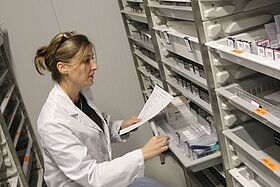 Une femme en blouse blanche cherche des médicaments dans des rayonnages pour préparer une commande d'après la liste qu'elle tient à la main