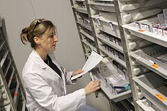 Une femme en blouse blanche cherche des médicaments dans des rayonnages pour préparer une commande d'après la liste qu'elle tient à la main