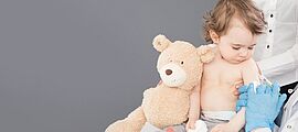 un très jeune enfant tenant un nounours contre lui regarde la piqûre qui s'enfonce dans son bras. Cette piqûre est tenue par des mains gantées en latex bleu dont on devine qu'elles appartiennent à un soignant ou un médecin. Derrière l'enfant, se tient une personne en civil qui tient l'enfant, certainement sa mère. On ne voit le visage que de l'enfant. - Enlarge picture (modal window)
