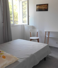 Chambre lumineuse avec un lit fait au premier plan, une fenêtre ouverte donnant sur des arbres, une chaise en bois et blanc, une console assortie et un tableau au mur.
